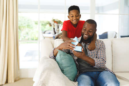 African american father opening gift from his son and smiling in living room. family spending time at home, father son relationship.の写真素材