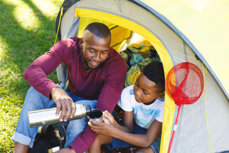 African american father with son having fun and sitting in tent pouring drinks in garden. family spending time at home.の写真素材