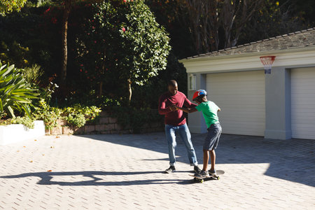 African american father smiling and helping son balancing on skateboard in garden. family spending time at home.の写真素材