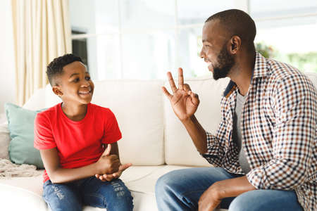 Happy african american father with son sitting on couch in living room talking sign language. father and son communicating without words.の写真素材