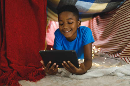 Happy african american boy lying in blanket fort, using tablet. spending time alone at home with technology.の写真素材