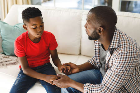 Serious african american father and son sitting on couch in living room talking and holding hands. family spending time at home, father son relationship.の写真素材