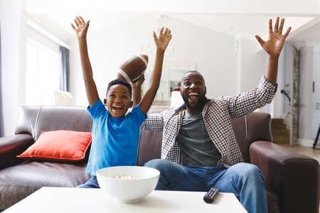 Excited african american father and son sitting on sofa, watching match on tv and cheering. family spending time together at home.の写真素材