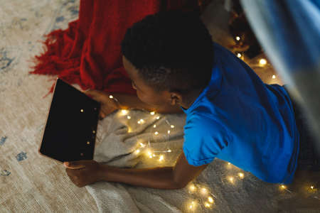 Happy african american boy lying in blanket fort, using tablet. spending time alone at home with...