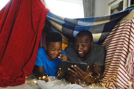 Happy african american father and son lying in blanket fort, using tablet. family spending time at home.の写真素材