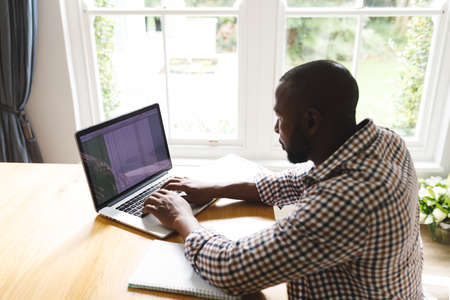 African american man sitting at table in dining room, working remotely using laptop. flexible...