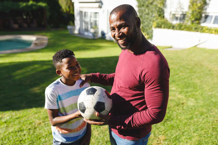 Portrait of smiling african american father with son embracing and holding football in sunny garden. family spending time at home.の写真素材