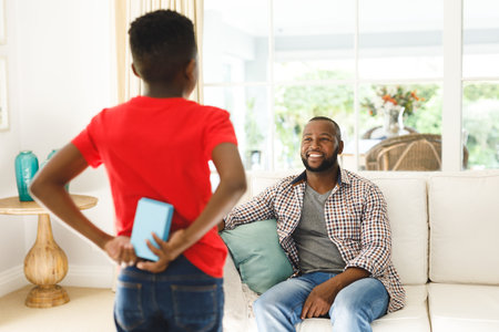 African american son giving gift his smiling father in living room. family spending time at home, father son relationship.の写真素材