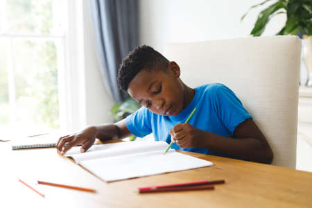African american boy sitting at table in living room, doing homework. spending time at home.