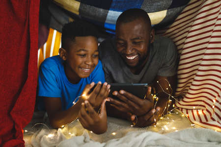 Happy african american father and son lying in blanket fort, using tablet. family spending time at...