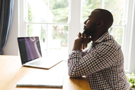 African american man sitting at table in dining room, working remotely using laptop. flexible working from home with technology.の写真素材