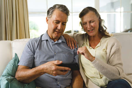 Happy senior caucasian couple in living room, sitting on sofa, using smartphone. retirement lifestyle, spending time at home.の写真素材