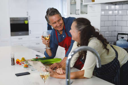 Happy senior caucasian couple in kitchen cooking together, drinking wine, using tablet. retirement...