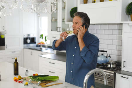 Senior caucasian man in modern kitchen, talking on smartphone and drinking wine. retirement lifestyle, spending time at home.の写真素材