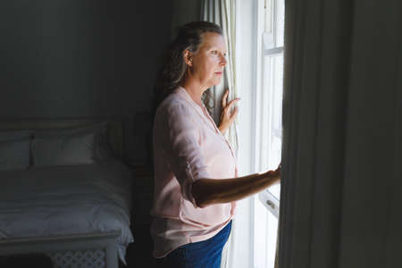 Thoughtful senior caucasian woman in bedroom, standing next to window, widening curtains. retirement...