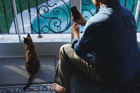 Senior caucasian man with cat in living room, sitting on sofa, using smartphone. retirement...