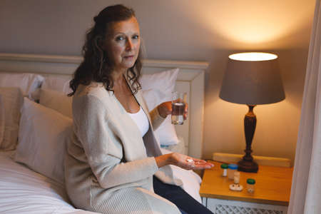 Portrait of senior caucasian woman in bedroom sitting on bed, holding pills. retirement lifestyle,...