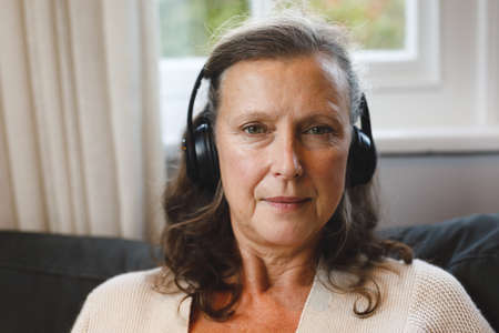Portrait of happy senior caucasian woman in living room sitting on sofa, wearing headphones....