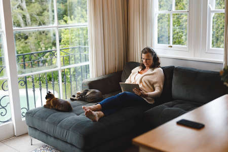 Happy senior caucasian woman in living room sitting on sofa, wearing headphones, using tablet. retirement lifestyle, spending time alone at home with technology.の写真素材