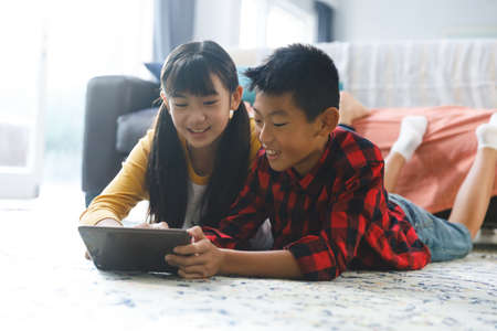 Smiling asian brother and sister lying in living room and using tablet. childhood leisure time at...