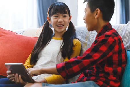 Smiling asian brother and sister sitting on couch and using tablet. childhood leisure time at home...