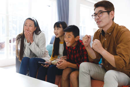 Smiling asian parents with son and daughter sitting on couch watching tv with popcorn. family...