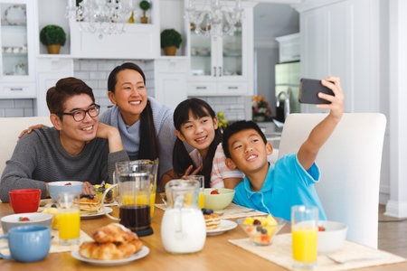 Smiling asian parents with son and daughter sitting at breakfast table taking selfie. family enjoying mealtime together at home.の写真素材