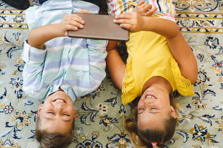 Smiling caucasian brother and sister lying in living room and using tablet. childhood leisure time,...
