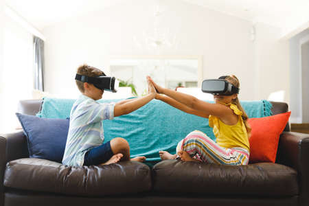 Caucasian brother and sister sitting on couch and using vr headsets in living room. childhood...