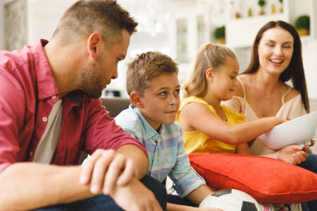 Happy caucasian parents, daughter and son sitting on couch talking and watching football match in...