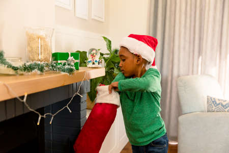 African american boy wearing santa hat and searching for present in christmas stocking. childhood, christmas, festivity and tradition at home.の写真素材