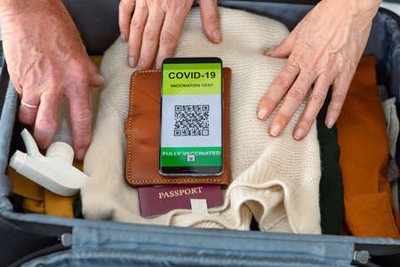 Hands of caucasian senior couple packing smartphone with covid vaccine passport into suitcase....