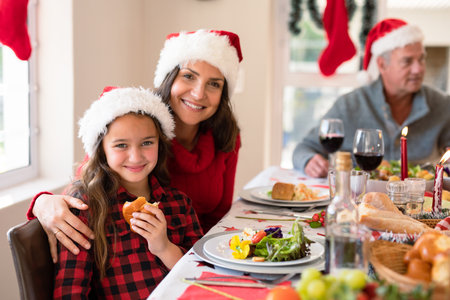 Happy caucasian mother and daughter wearing santa hats looking at camera at christmas time. family christmas time and festivity together at home.の写真素材