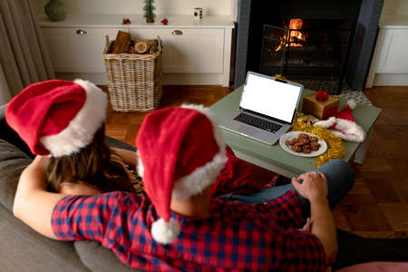 Back view of caucasian couple wearing santa hats, using laptop with copy space at christmas time. christmas, festivity and communication technology.の写真素材