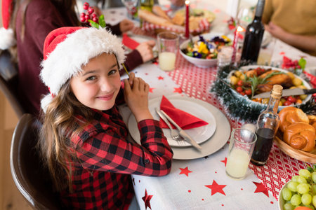 Happy caucasian girl wearing santa hat sitting at christmas table and looking at camera. family christmas time and festivity together at home.の写真素材