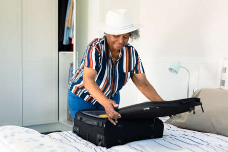 African american senior woman wearing hat packing suitcase in bedroom. travel preparation during covid 19 pandemic.の写真素材