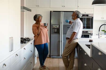 Happy african american senior couple standing in kitchen, holding mugs with coffee and talking. healthy retirement lifestyle at home.の写真素材