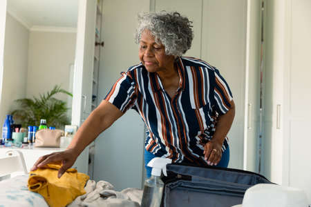 Focused african american senior woman packing suitcase in bedroom. travel preparation during covid 19 pandemic.の写真素材