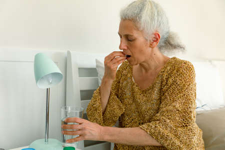 Tired caucasian senior woman taking pills and sitting on bed. senior health and lifestyle at home.の写真素材