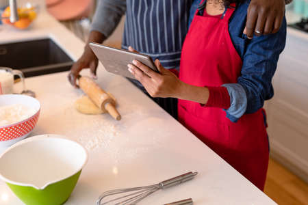Midsection of african american couple wearing aprons, baking together and using tablet. family time, having fun together at home.の写真素材