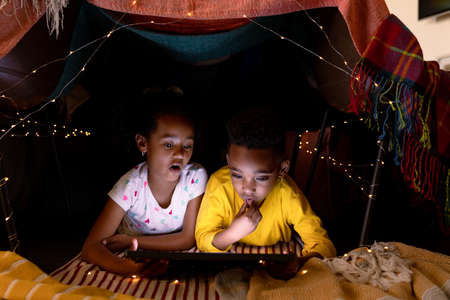 Surprised african american siblings lying in makeshift tent, using tablet. childhood, christmas, festivity and tradition at home.の写真素材