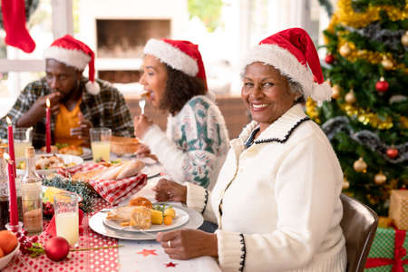 Happy african american grandmother wearing santa hat sitting at christmas table with family. family christmas time and festivity together at home.の写真素材