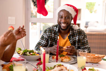 Happy african american man wearing santa hat sitting at christmas table. family christmas time and festivity together at home.の写真素材