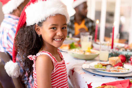 Happy african american girl wearing santa hat sitting at christmas table. family christmas time and festivity together at home.の写真素材