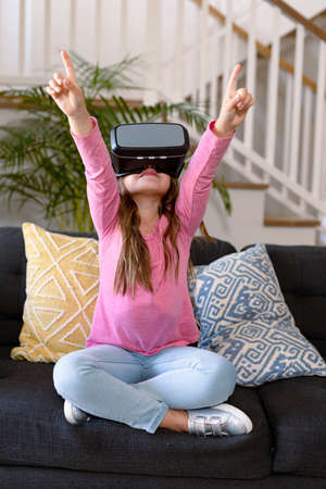 Happy caucasian girl sitting on sofa, wearing vr headset, having fun. childhood, leisure and discovery using technology at home.の写真素材