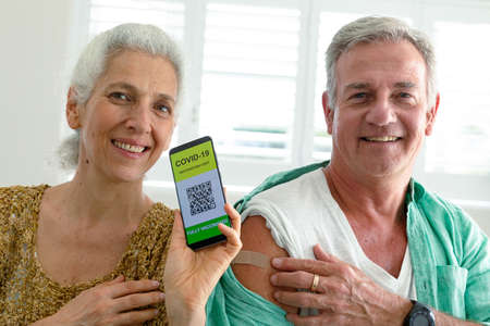 Smiling caucasian senior couple showing smartphone with covid 19 vaccine passport on screen. senior health and lifestyle during covid 19 pandemic.の写真素材