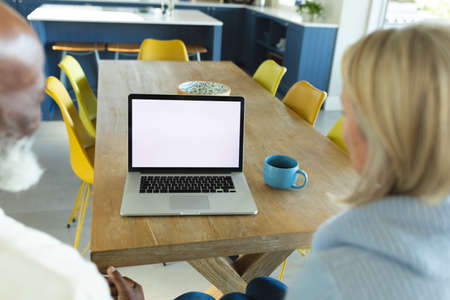 Happy senior diverse couple in kitchen sitting at table, using laptop. retirement lifestyle, at home with technology.の写真素材