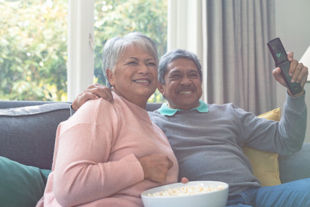 Two diverse senior couple sitting on sofa, watching tv and having fun. retirement lifestyle relaxing at home with technology.の写真素材