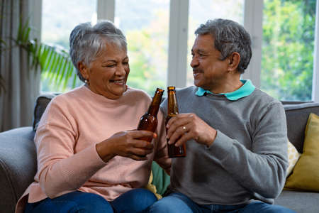 Happy diverse senior couple sitting on sofa and drinking beer. retirement lifestyle and relaxing at home.の写真素材
