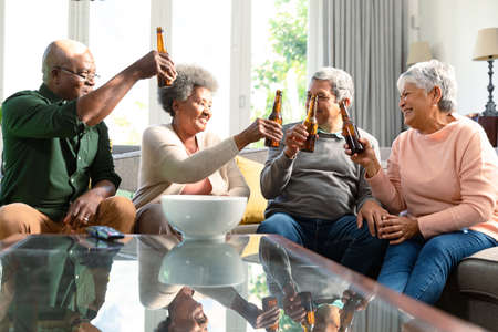 Two diverse senior couples sitting on sofa with beer and having fun. retirement lifestyle relaxing at home with technology.の写真素材